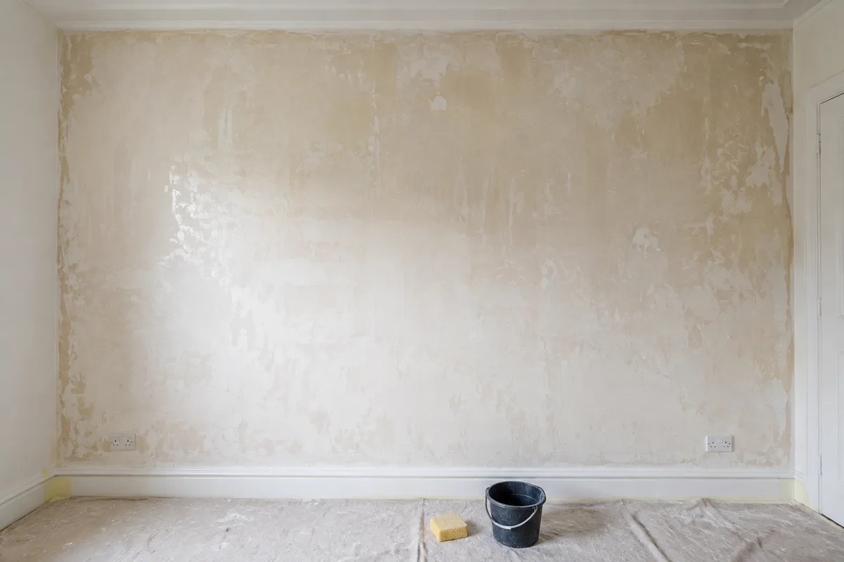 Mur de chambre entièrement dépouillé de son papier peint, traces de colle visibles, plinthes protégées